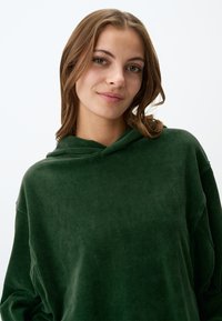 Felpa verde in velluto a coste con superficie testurizzata, dotata di cappuccio e spalle scese. Design semplice, senza motivi visibili o accessori.