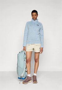 Ανοιχτό μπλε fleece πουλόβερ με φερμουάρ μέχρι τη μέση, μπεζ σορτς, καφέ παπούτσια πεζοπορίας και ανοιχτό μπλε σακίδιο πλάτης. Λεπτή υφή, casual σχεδιασμός.