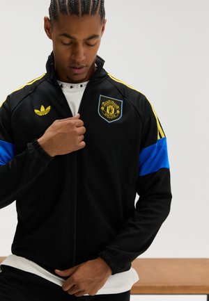 Casaco preto com fecho de correr, com detalhes amarelos e azuis, apresentando o emblema do Manchester United e o logótipo da Adidas no peito e nas mangas.