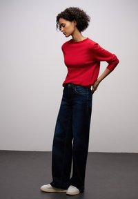 Czerwony sweter z dzianiny z długimi rękawami, zestawiony z ciemnoniebieskimi szerokimi dżinsami. Modelka nosi białe trampki. Neutralne tło.