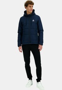 Giacca imbottita blu navy con cappuccio, dotata di chiusura con zip e logo sul petto, abbinata a pantaloni neri e sneakers bianche.