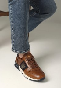 Hnědé kožené tenisky s černými akcenty, zaoblenou špičkou a šněrovacím designem. Obsahují značku na jazyku a texturovanou gumovou podrážku.