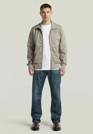 Junger Mann, der eine hellgraue Jacke mit Reißverschluss über einem weißen Hemd, blaue Jeans und dunkelbraune Schuhe trägt, vor einem schlichten Hintergrund steht.