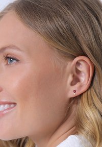 Boucles d'oreilles en or avec une pierre précieuse rouge en forme de cœur et une plus petite pierre claire, mises en valeur sur une peau claire et des cheveux blonds ondulés.