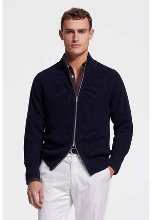 Man in een marineblauwe trui met rits over een donker shirt en een witte broek, staande tegen een effen lichte achtergrond.