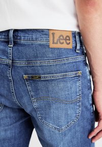 Jeans de mezclilla azul con una etiqueta de cuero con el logo "Lee" en la parte trasera, costuras amarillas y un diseño clásico de cinco bolsillos.
