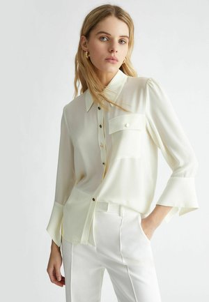 Camicia button-up di colore crema chiaro realizzata in tessuto setoso, con taschino sul petto, bottoni neri e polsini svasati. Abbinata a pantaloni bianchi.