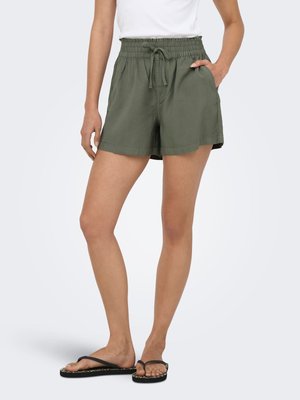 Mujer con pantalones cortos verdes oliva con cordón y cintura elástica, camiseta blanca y chanclas negras, de pie con una mano en el bolsillo.