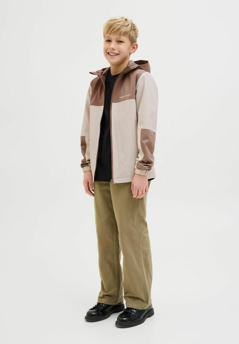 Veste légère, beige et marron à blocs de couleur avec capuche, t-shirt noir, pantalons verts olive et chaussures noires, avec des poignets élastiques.