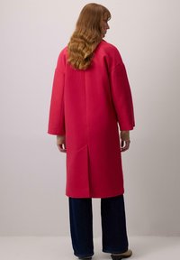 Hosszú rózsaszín kabát laza szabással, esett vállakkal és hátsó szellőző résszel. Az anyag simának és puhanak tűnik. Széles lábú sötét nadrággal párosítva.