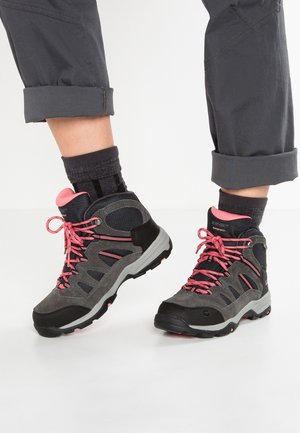 Szare i czarne buty trekkingowe z różowymi sznurówkami i akcentami, noszone z czarnymi skarpetkami i podwiniętymi szarymi spodniami na białym tle.
