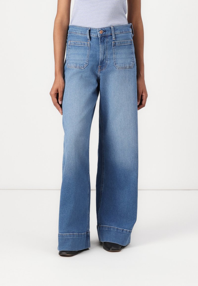 Vaqueros de denim azul de pierna ancha con cintura alta, cinco bolsillos y costuras oscuras. La tela parece resistente y tiene una textura suave.