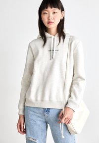 Lichtgrijze hoodie met koordjes, voorzien van het logo "Calvin Klein Jeans". Combinatie met blauwe distressed jeans en een crèmekleurige schoudertas.