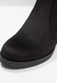 Gros plan d'une botte cheville en daim noir avec un bout arrondi et une semelle en caoutchouc légèrement texturée sur fond blanc.