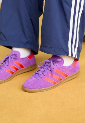 Baskets en suede violet avec des rayures et des accents orange, présentant une semelle texturée et des lacets plats. Portées avec un pantalon de jogging bleu marine.