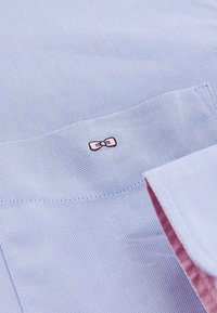 Tissu texturé bleu clair avec un petit nœud rose brodé sur la poche poitrine ; doublure rose contrastante visible au bord du col.