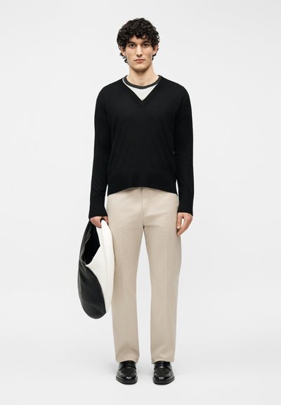 Άνδρας με σγουρά μαλλιά που φοράει μαύρο πουλόβερ με V λαιμόκοψη, μπεζ παντελόνια, μαύρα παπούτσια, κρατώντας μια μεγάλη ασπρόμαυρη τσάντα σε απλό φόντο.