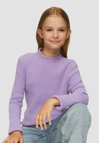 Lila gestrickter Pullover mit hohem Kragen, Rippstruktur und langen Ärmeln. Getragen mit hellblauen Jeans.