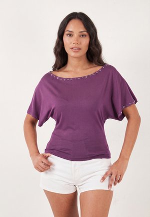 Mujer con cabello largo y oscuro, vistiendo una camiseta morada de manga corta con detalles de ojales y pantalones cortos blancos, de pie con las manos en las caderas.