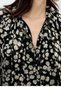 Blouse noire avec motif floral de marguerites blanches et jaunes, col en V, et boutons à l'avant, portée par une personne aux longs cheveux bruns.