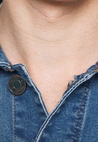 Närbild av halsen och kragen på en blå denimjacka med synlig knapp och fransad kant på högra sidan.