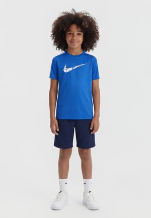 Barn med krøllete hår iført blå Nike T-skjorte, marineblå shorts, hvite Nike-sokker og hvite joggesko, stående mot en enkel lys bakgrunn.