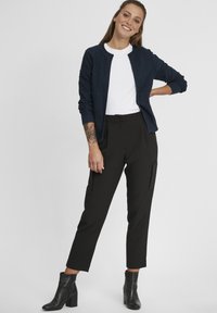 Cardigan texturé bleu marine sur un t-shirt blanc, associé à un pantalon noir fuselé avec poches latérales et ourlet à la cheville, complété par des bottes à talons noires.