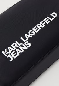 Juodas nailono maišelis su iškilniu baltu logotipu "KARL LAGERFELD JEANS", lygia tekstūra, stačiakampio formos, be matomų detalių ar papildomų elementų.
