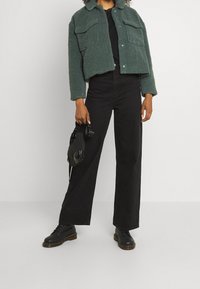 Grøn tekstureret jakke med to forlommer over en sort top. Sorte jeans med vide ben og sorte snørestøvler fuldender outfittet. Holder sko.