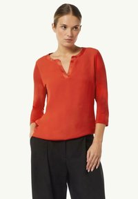 Rote, halbtransparente Bluse mit V-Ausschnitt und Dreiviertelärmeln. Hat eine glatte Textur und leichten Glanz, kombiniert mit schwarzen Hosen.