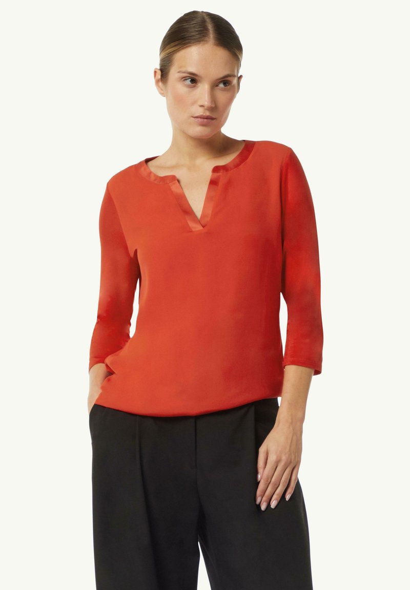Rote, halbtransparente Bluse mit V-Ausschnitt und Dreiviertelärmeln. Hat eine glatte Textur und leichten Glanz, kombiniert mit schwarzen Hosen.