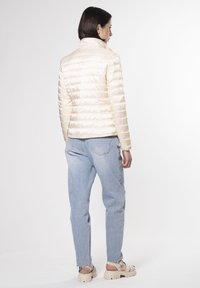 Giacca puffa di colore crema chiaro con colletto alto, texture trapuntata e forma aderente. Abbinata a jeans azzurri chiari e sandali chunky.