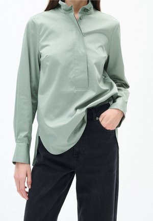Vrouw draagt een lichtgroene blouse met lange mouwen en knopen, ingestopt in hoog getailleerde zwarte jeans, met een hand in de zak.