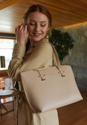 Beige leather tote bag with gold hardware, featuring two top handles and a smooth, structured design. Simple logo detail on the front.