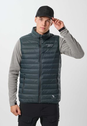 Jeune homme portant une casquette noire, une chemise grise à manches longues avec des motifs, et une veste matelassée vert foncé avec des poches zippées.