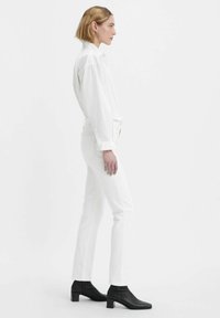 Jeans in denim bianco con vestibilità slim e cuciture a contrasto, abbinati a una camicia bianca con bottoni e stivaletti neri.