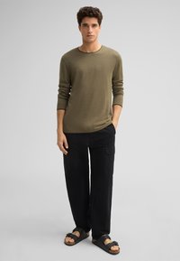 Olivenfarbendes Langarmhemd mit Rundhalsausschnitt, kombiniert mit lockeren schwarzen Cargo-Hosen und schwarzen Sandalen mit Riemen. Glatte Stofftextur.