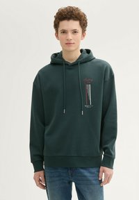 Ciemnozielona bluza z kapturem z nadrukiem na przodzie, sznurkami zakończonymi metalowymi końcówkami oraz ściągaczami przy mankietach i u dołu. Noszona z niebieskimi dżinsami.