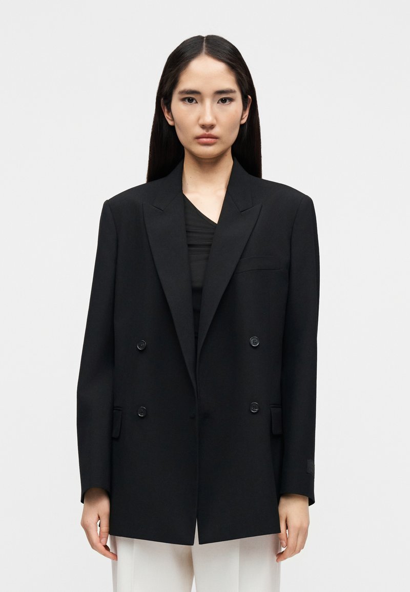 Veste noire à double boutonnage en tissu structuré, dotée de grands boutons, de revers crantés et de poches latérales. Portée sur un haut noir.