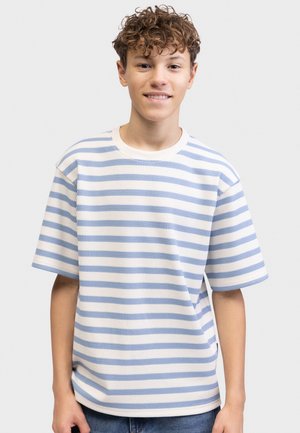 Έφηβος με μπούκλες που χαμογελά, φορώντας μια λευκή και γαλάζια κοντομάνικη μπλούζα με οριζόντιες ρίγες, στέκεται μπροστά σε ένα μονόχρωμο φόντο.