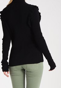 Svart ribbad tröja med volangdetaljer på axlarna, med långa ärmar och en figurnära design, matchad med gröna figurnära byxor.