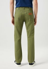 Mężczyzna noszący oliwkowe dżinsy Wrangler i czarne trampki, stojący tyłem do kamery na jednolitym tle.