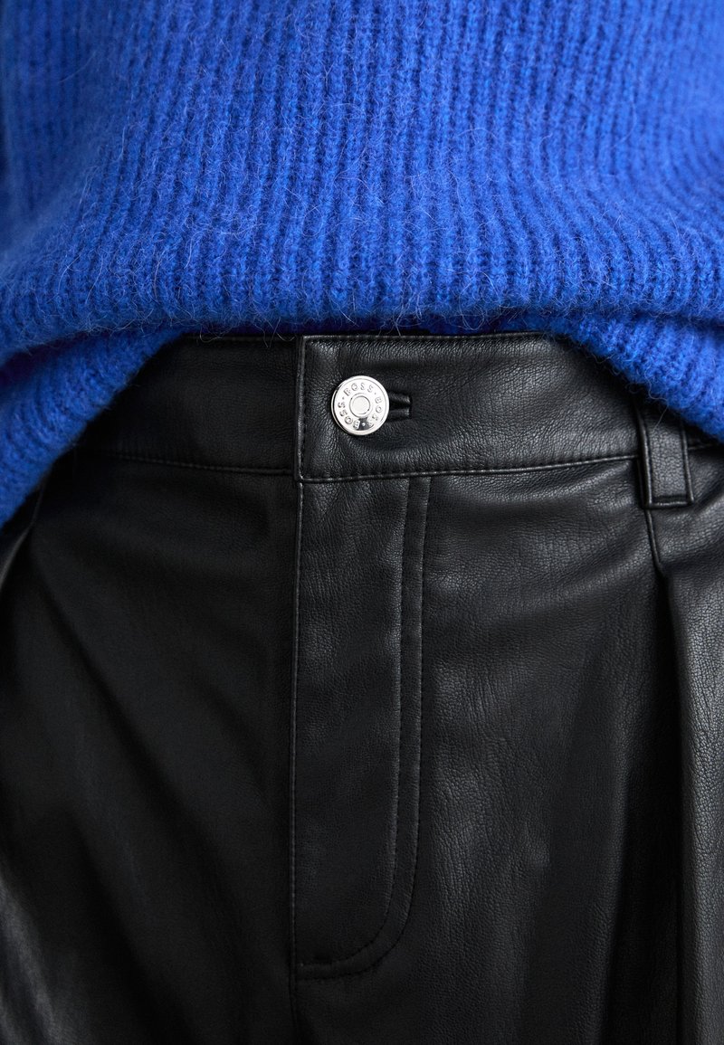 Pull en maille côtelée bleu sur pantalons en cuir noir, avec une fermeture à bouton en argent avec détail gravé et texture lisse.