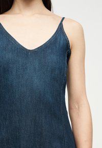 Γυναίκα που φοράει σκούρο μπλε φόρεμα από τζιν χωρίς μανίκια με λεπτούς ιμάντες στους ώμους και V-λαιμόκοψη σε ουδέτερο φόντο.