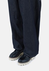 Pantalones de mezclilla de pierna ancha en azul oscuro con detalle de cremallera, combinados con zapatos de cuero negro con una suela gruesa en crema y costuras destacadas.