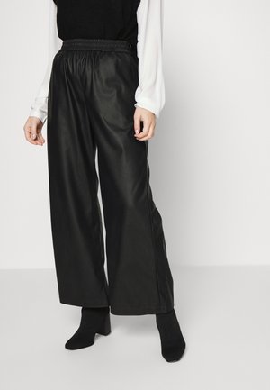 Vrouw draagt zwarte wijde leren broek, zwarte laarzen met hak en een zwart-wit top met lange mouwen tegen een effen achtergrond.