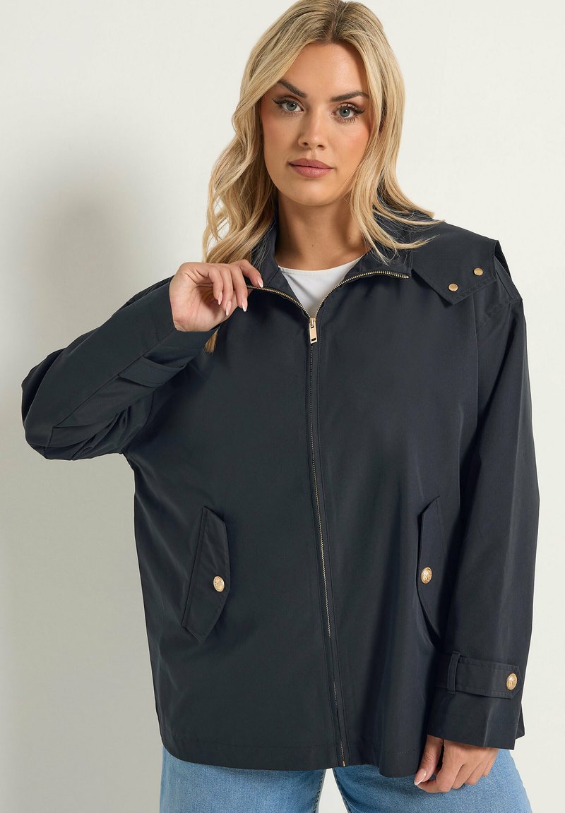 Femme blonde portant une veste noire surdimensionnée avec des poches avant et des boutons dorés, tenant la fermeture éclair près du col, sur un fond neutre.