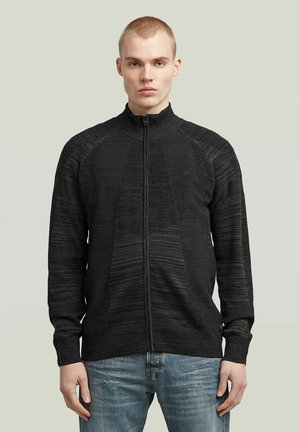 Jeune homme portant un pull en maille noir zippé et un jean bleu, debout devant un fond uni gris clair.