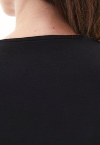 Parte posteriore del collo e della spalla di una persona con capelli castani, che indossa un top nero a girocollo su uno sfondo bianco.