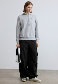 Hall halli puuvillane kapuutsiga dressipluus, millel on pael ja väike roheline logo. Kombineeritud mustade kaubapükstega ja hõbedaste tennistega, käes metallik värvi kott.